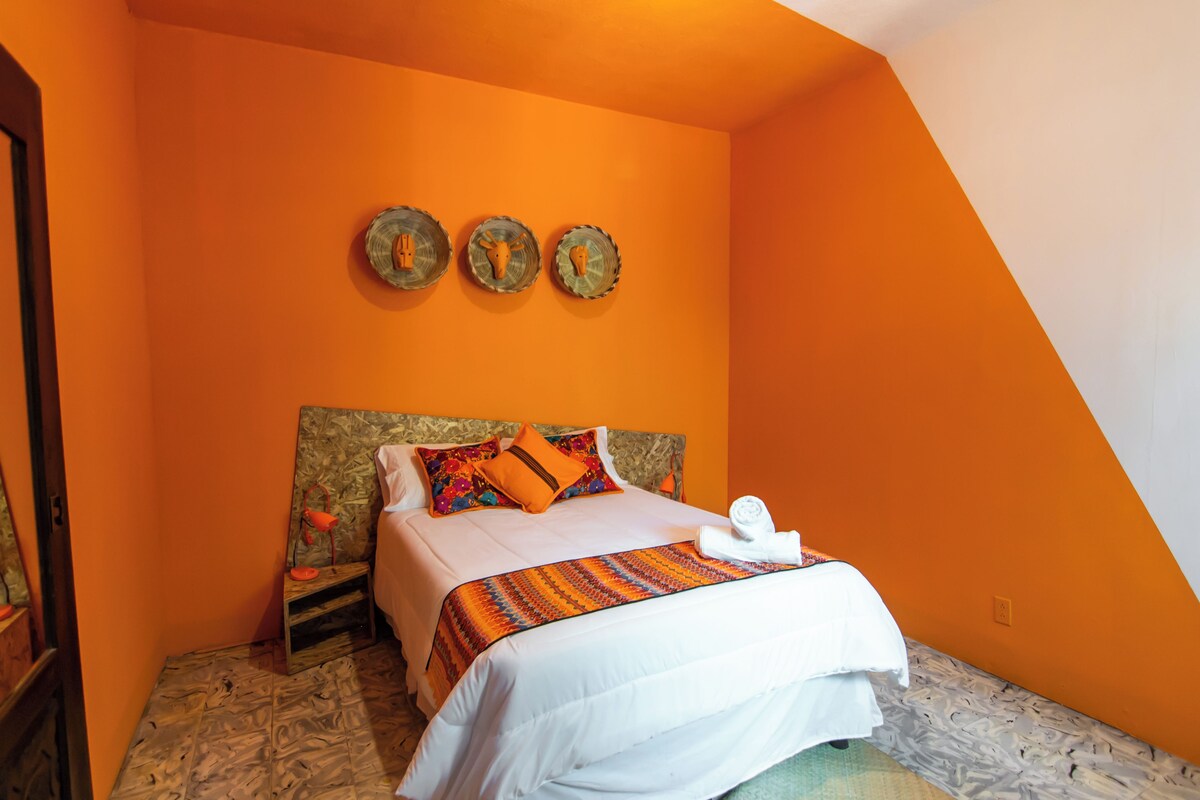 A bedroom with an orange wall, a bed with white bedding, a wooden headboard, and three wall-mounted decorative items.