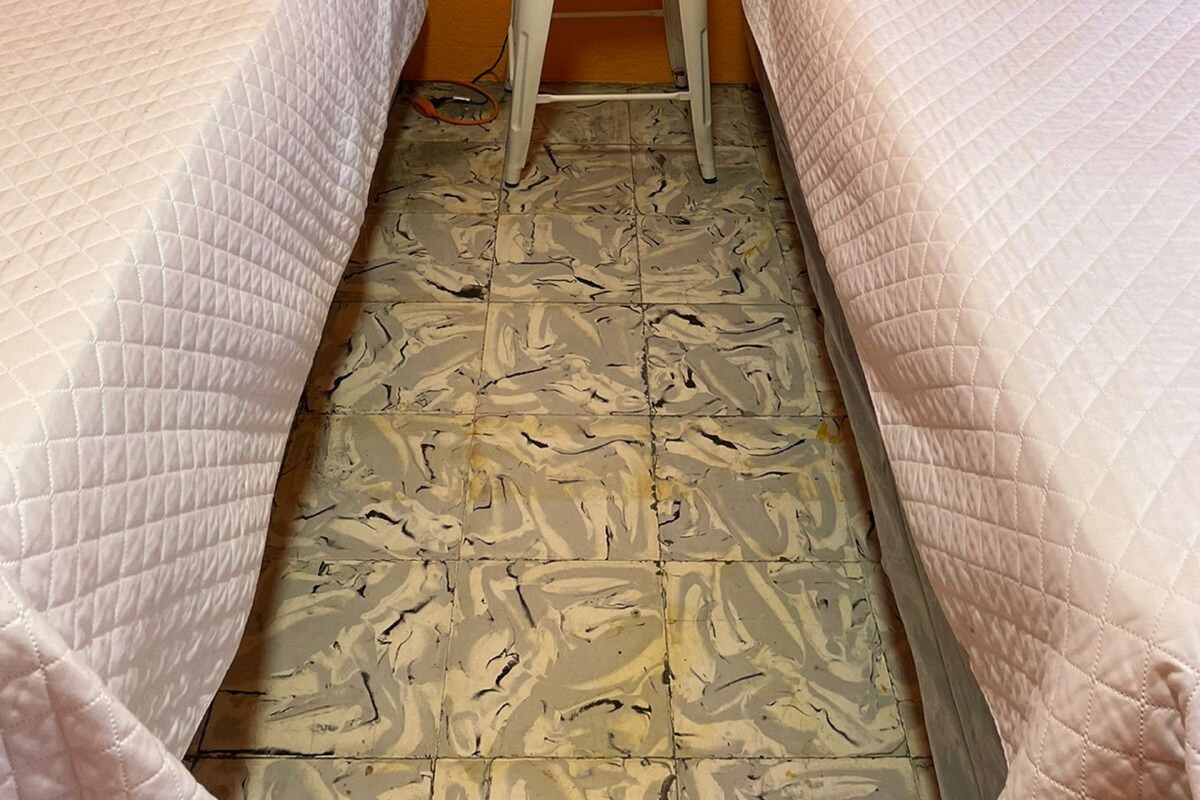 A hotel room with two beds, each with a white quilted bedspread and a red pillow. There is a small white nightstand with an orange lamp between the beds. The floor has a patterned tile design.