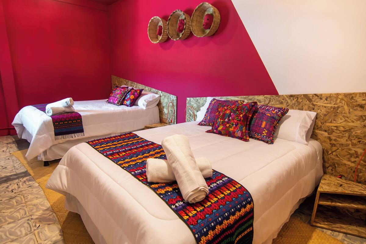 A hotel room with two beds, each with white linens and colorful blankets. There are decorative pillows and a woven wall art piece above the beds.