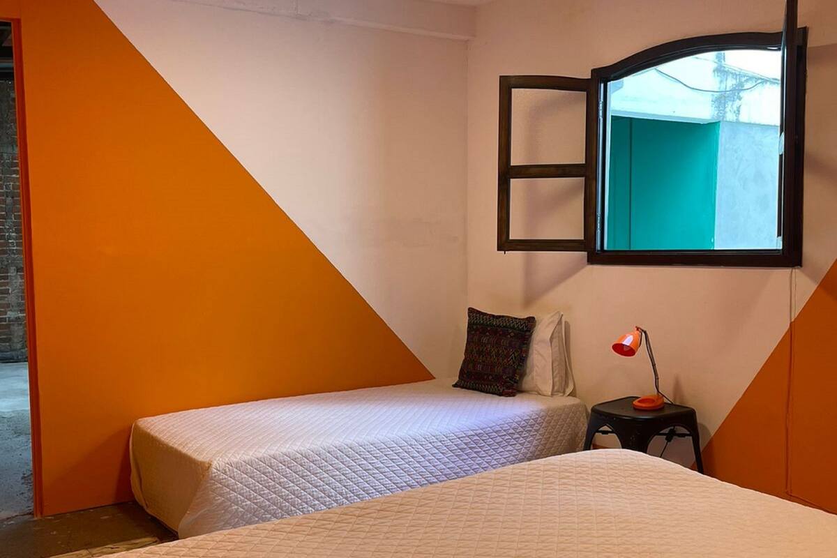 A bedroom with two beds, a small table with a lamp, and a window with a view of the outside.