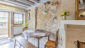 A rustic dining area with a wooden table and chairs, a stone wall, and a window with a view of greenery. - Le Verger - Paris L'hopital (PARIS L'HOPITAL)