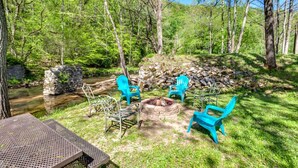 A patio area with a fire pit, blue chairs, and a view of a forested area with a stream. - Calm Country Oasis - Creek, ATV Trls & Norris Lake (Speedwell)