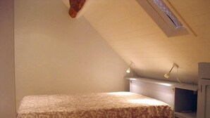 A small, single-bed room with a sloped ceiling and a skylight. There is a bedside table with a lamp and a small shelf.