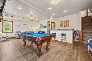 A pool table in a room with a bar and stools, a staircase, and a bookshelf.