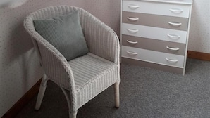 Een hoekappartement met een witte rieten stoel, een witte dressoir met lades, een lamp, en een schilderij van een roze bloem aan de muur.