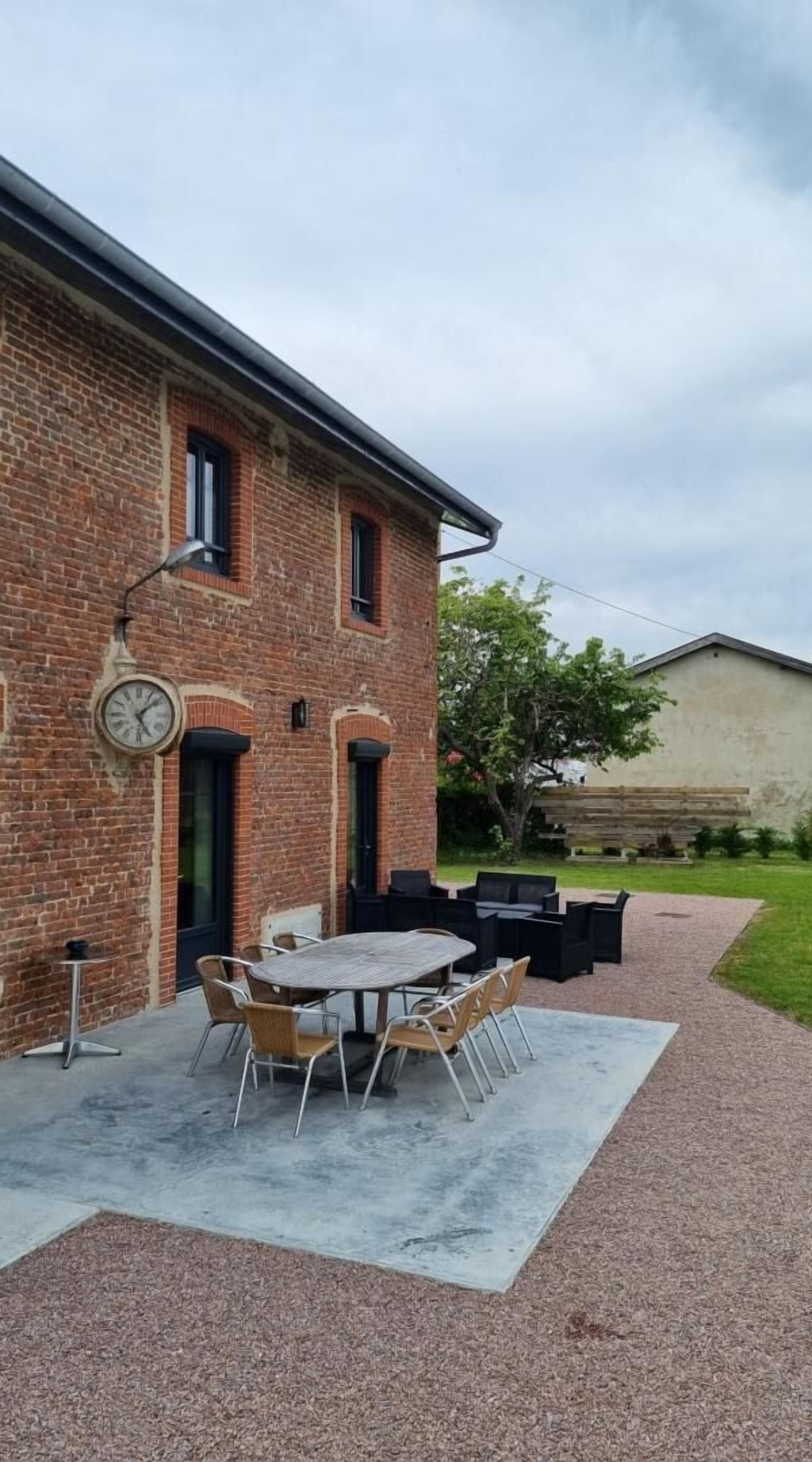 Edificio de ladrillo con un patio que tiene una mesa redonda y sillas, un reloj y asientos al aire libre.