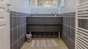 Ein schmales Badezimmer mit Fliesenboden, einem Waschbecken mit Spiegel darüber und einem Handtuchhalter rechts.