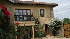 A two-story stone house with a balcony, a gravel driveway, and flowering plants.