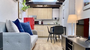 Un espace de vie compact comprenant un canapé gris, une table à manger avec des chaises, un coin cuisine avec des meubles en bois et un téléviseur posé sur un meuble.