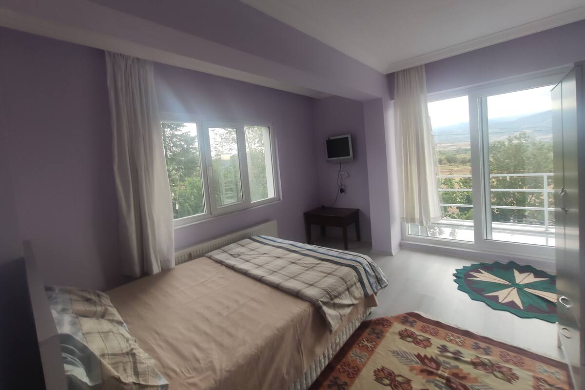 A bedroom with a bed, a TV, a small table, and large windows with curtains.