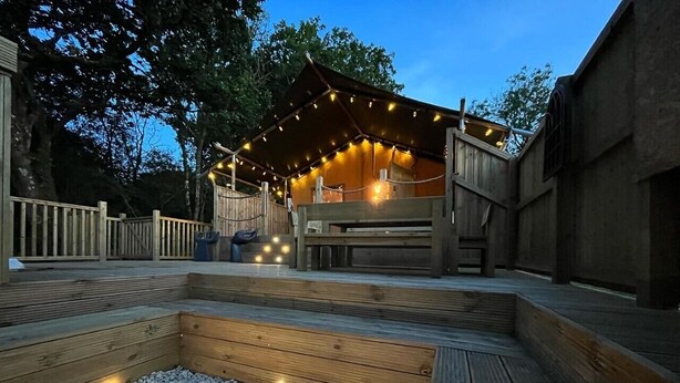 Une terrasse extérieure en bois, dotée d’un foyer, et entourée d’une clôture en bois et d’arbres.