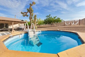 A swimming pool with a wooden deck, a covered seating area, and a fenced yard with trees.