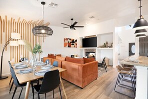 A modern living room with a sofa, dining table, and chairs. There is a fireplace, a television, and a ceiling fan. - Alana House-Hot Tub, Pool, Fire Pit & Near Trails (Joshua Tree)