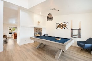 A pool table in a room with a fireplace, a hanging light fixture, and a wall-mounted rack of pool cues. - Alana House-Hot Tub, Pool, Fire Pit & Near Trails (Joshua Tree)