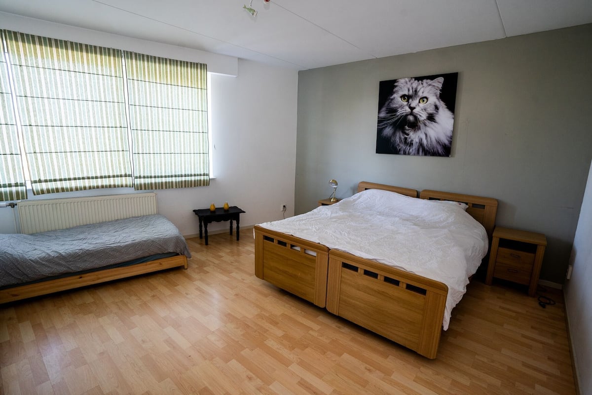 Une chambre à coucher avec un lit en bois, un plus petit lit, une table de chevet, une commode, un radiateur et une fenêtre avec des stores.
