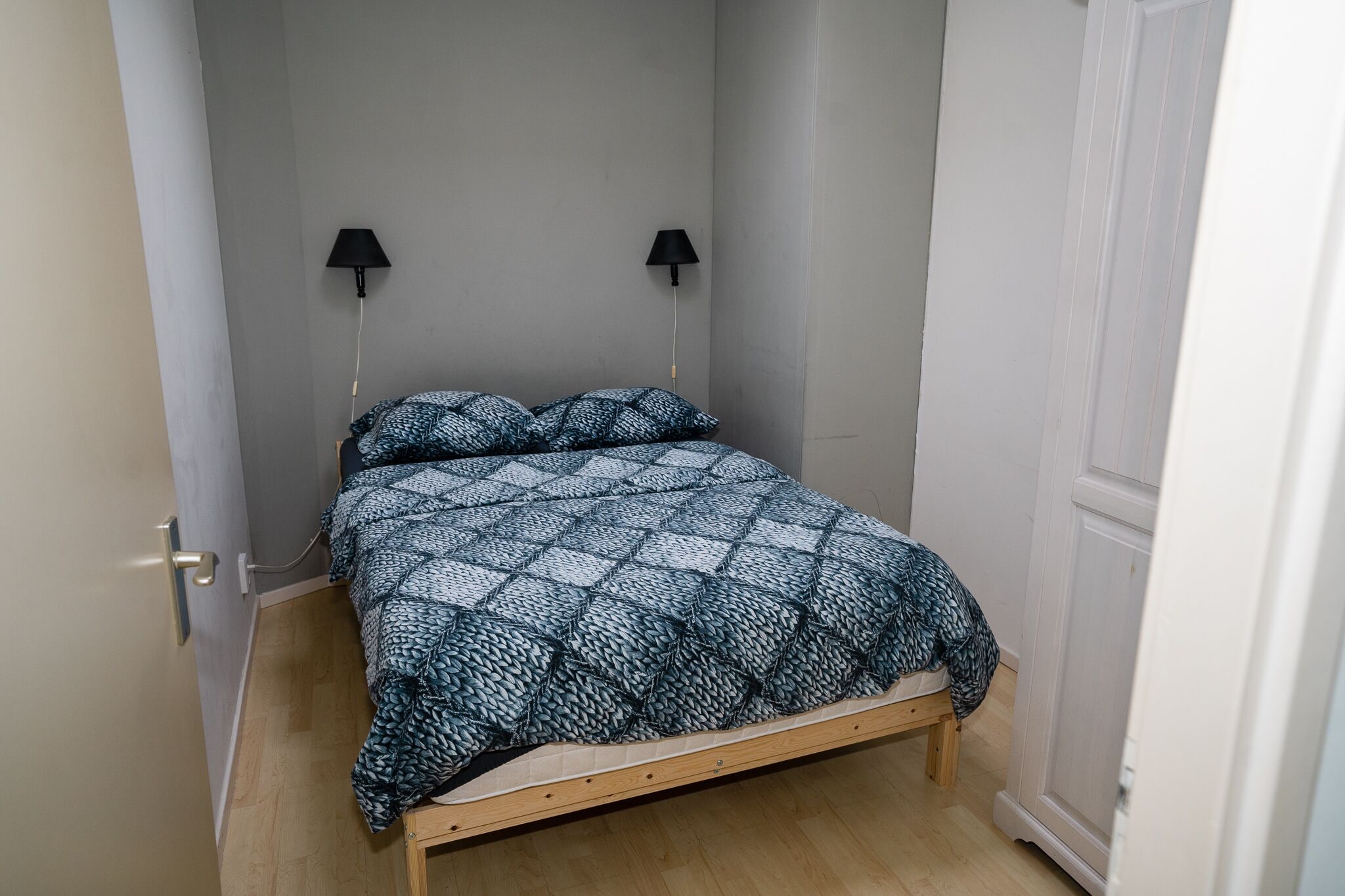 A small, narrow room with a bed, two wall-mounted lamps, and a door.