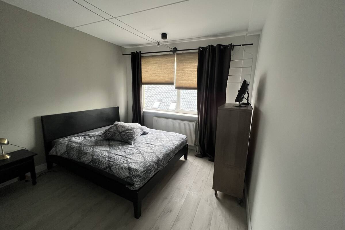Une chambre à coucher comprenant un lit, une table de chevet, une commode et une fenêtre avec des rideaux.
