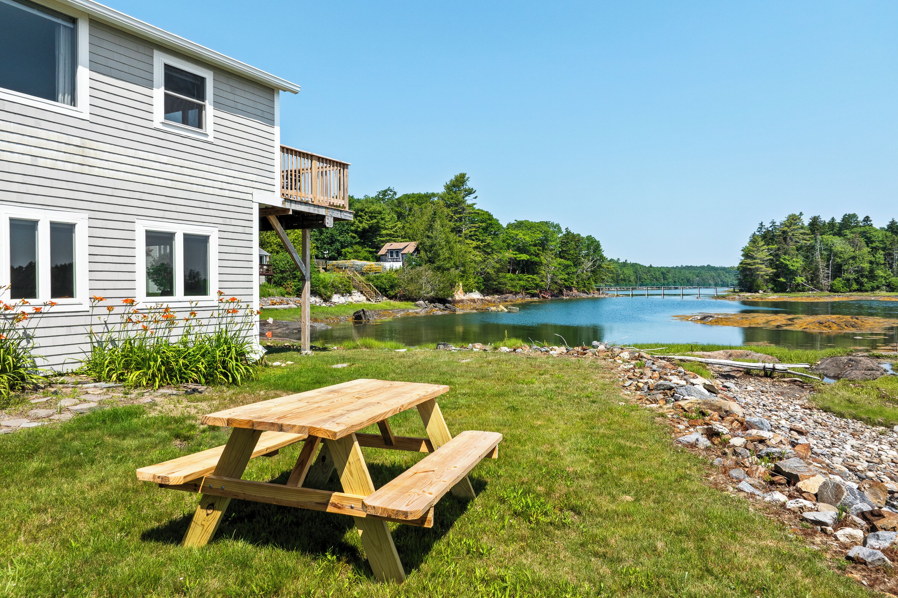 Ein Holztisch für ein Picknick in einer grünen Wiese, im Hintergrund ein See und ein Haus.