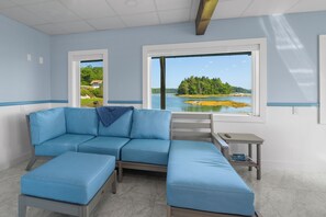 A room with a sectional sofa, a small table, and a view of a lake and forest. - Seal’s Haul. Water's edge living in this 1 BR and 1 BA first floor unit. (Harpswell)
