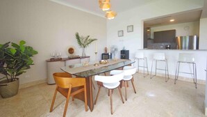 A modern dining area with a glass table, wooden chairs, and a kitchen visible in the background. - Stylish resort penthouse with amazing views (Punta Cana)