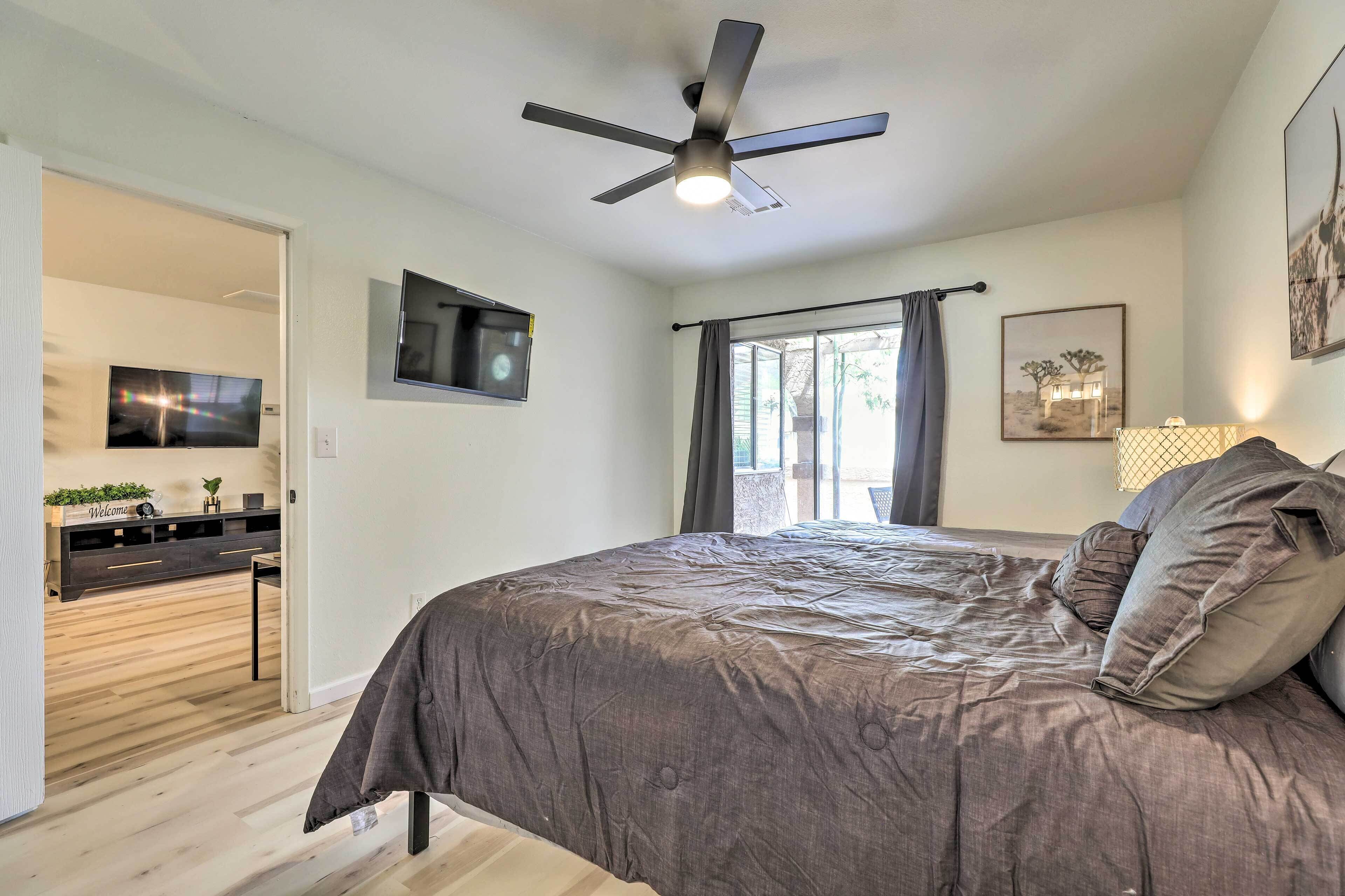 A bedroom with a bed, a ceiling fan, a TV, a window with curtains, and a desk with a plant.
