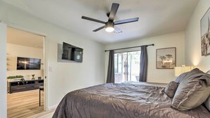 Une chambre à coucher avec un lit, un ventilateur de plafond, une télévision, une fenêtre avec des rideaux et un bureau avec une plante.