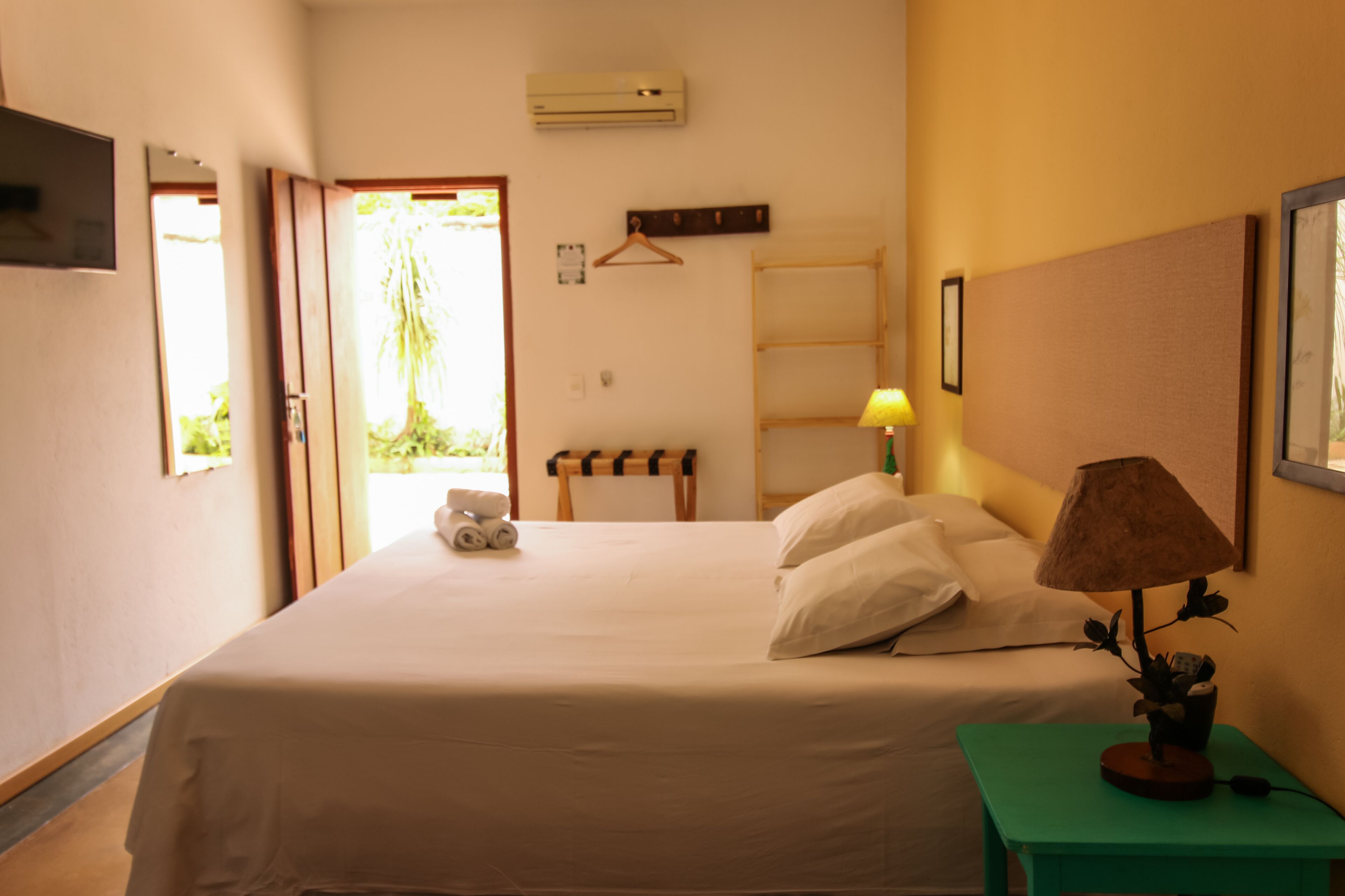 A hotel room with a bed, a bedside table with a lamp, a wall-mounted TV, and a window with a view of greenery.