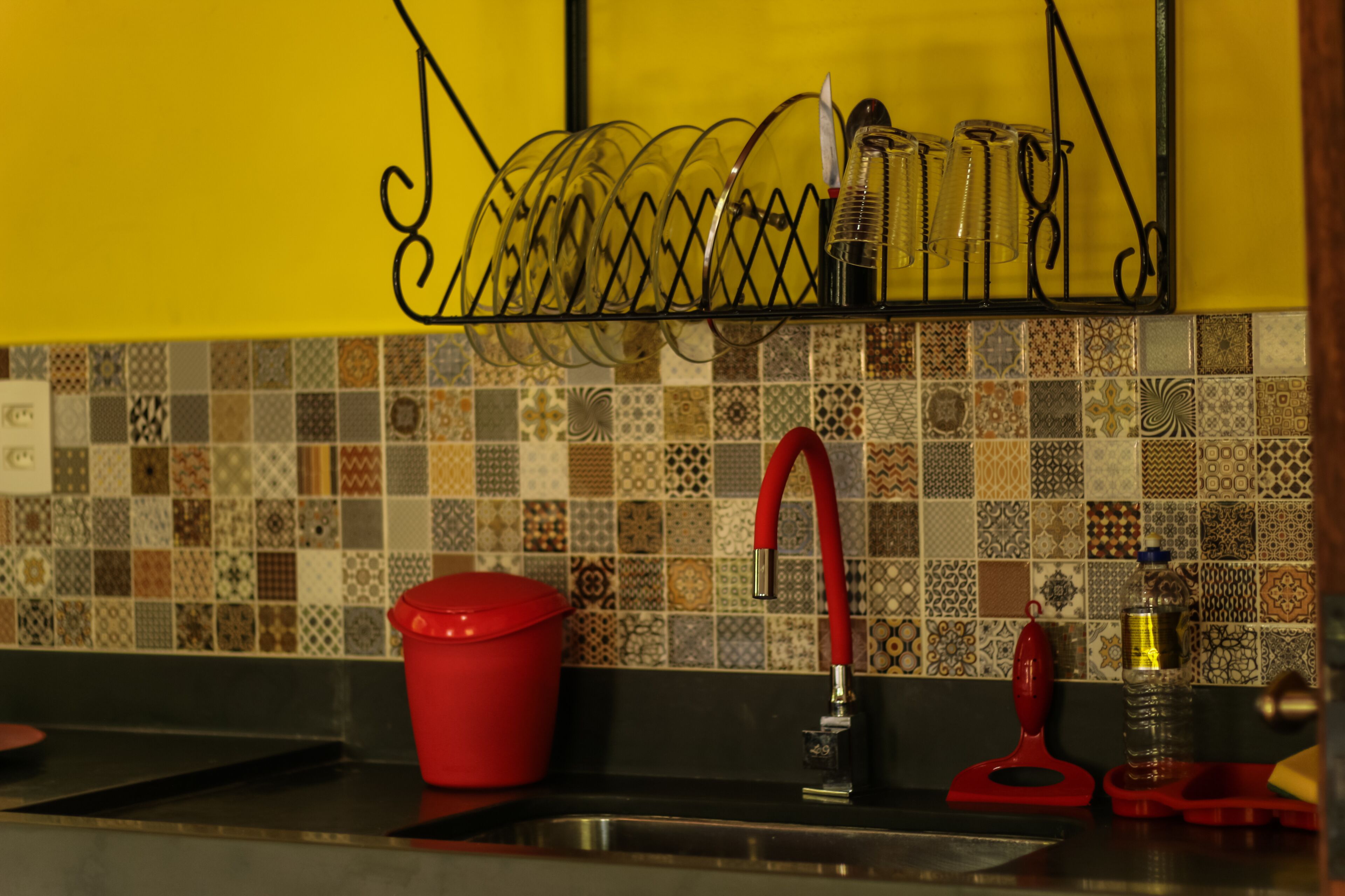 A kitchen with a tiled backsplash, a red bucket, a red faucet, and a metal dish rack.