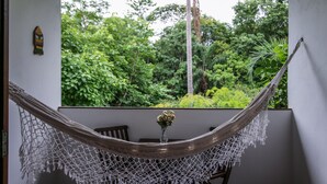 A hammock with a macrame cover, a small table with a vase of flowers, and a view of lush greenery through a window. - Pousada Arvoredo (Pirenópolis)