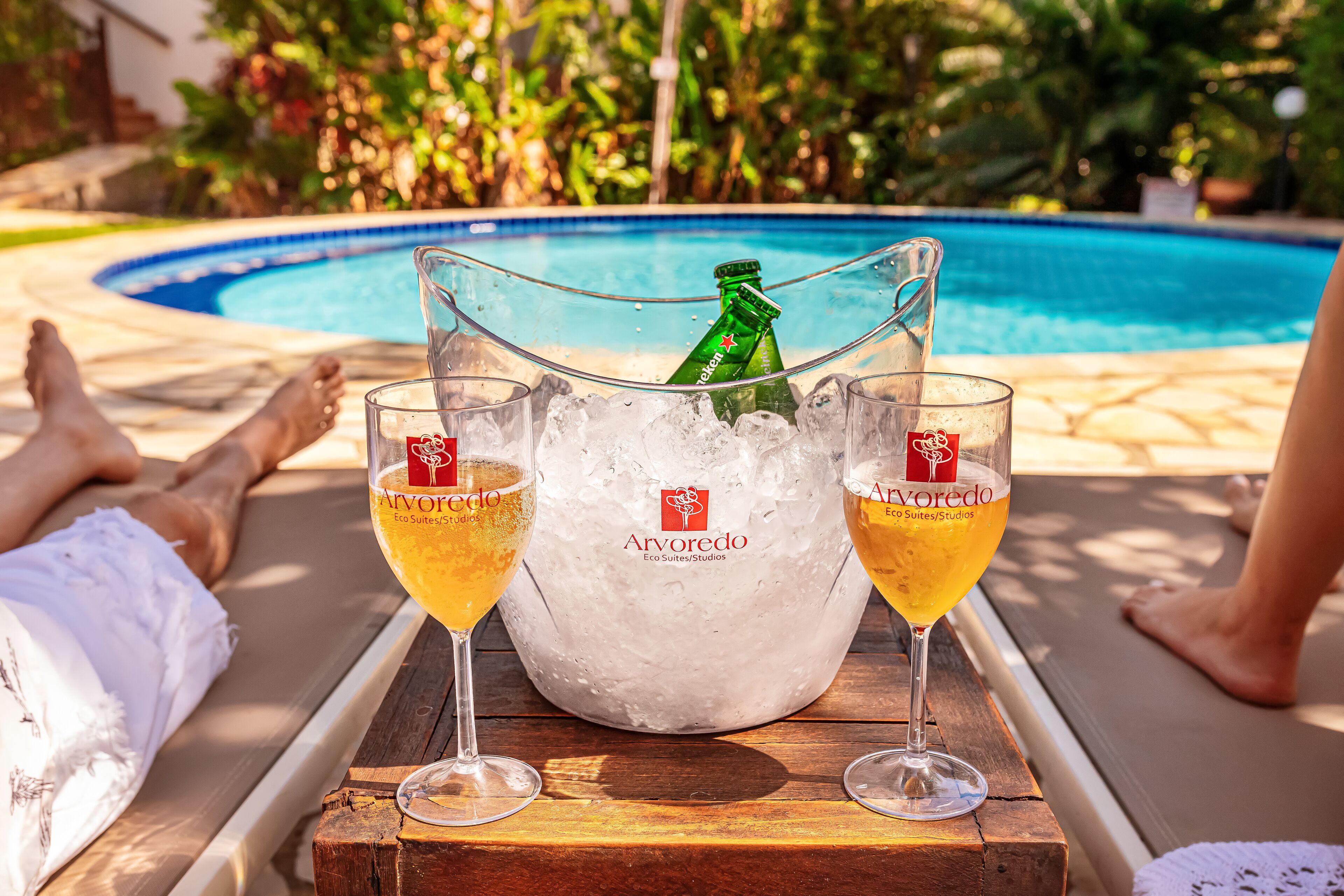 A poolside setting with a wooden table holding two glasses of a yellow beverage and a bottle of beer in an ice bucket.