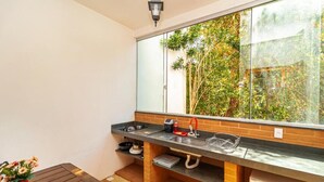 A compact kitchen with a large window, a sink, and a small dining area with a table and a potted plant. - Pousada Arvoredo (Pirenópolis)