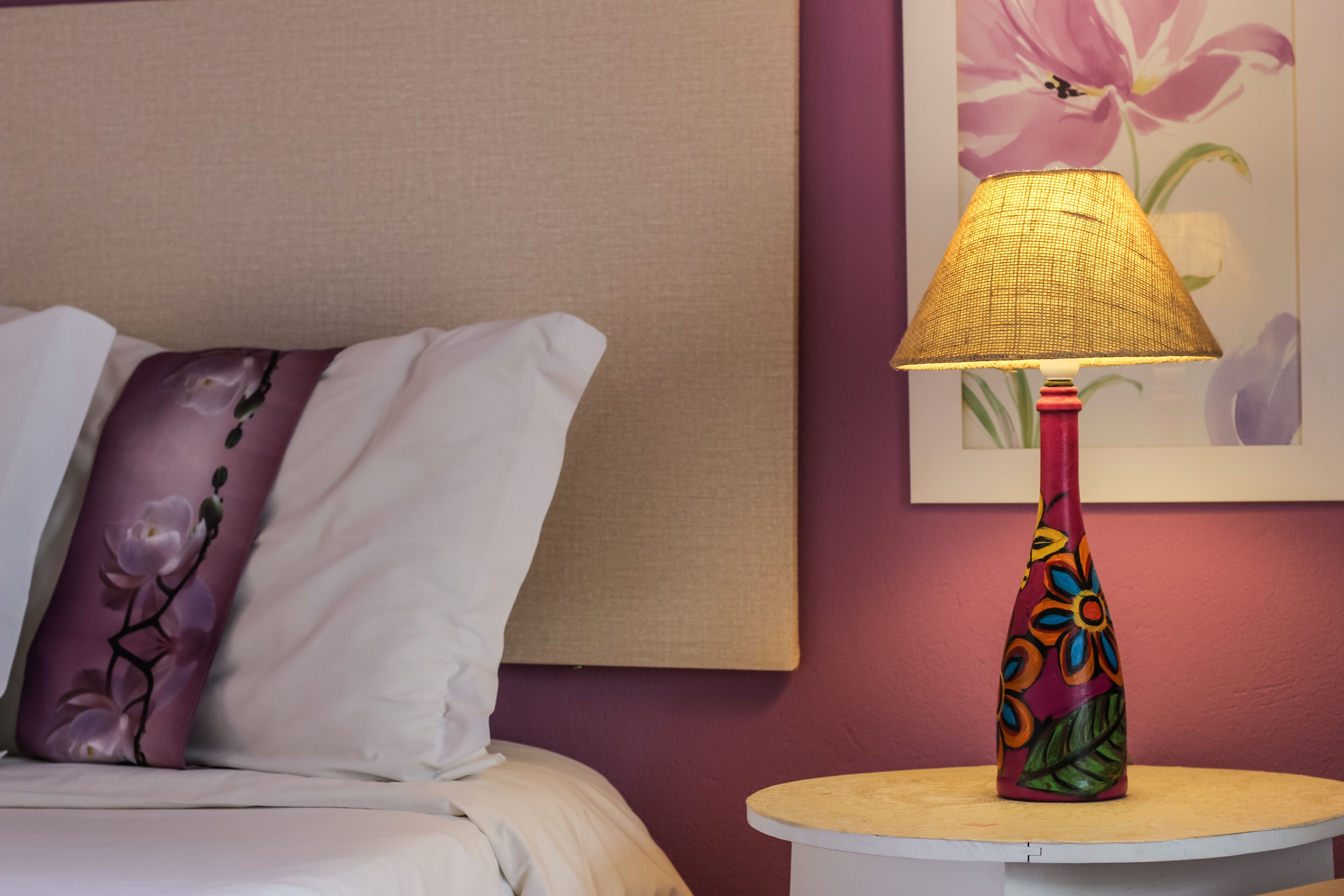 Ein Bett mit einem weißen und violetten Kissen, ein runder Tisch mit einer farbenfrohen Lampe und ein Blumenbild an der Wand.