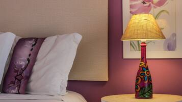 Ein Bett mit einem weißen und violetten Kissen, ein runder Tisch mit einer farbenfrohen Lampe und ein Blumenbild an der Wand.