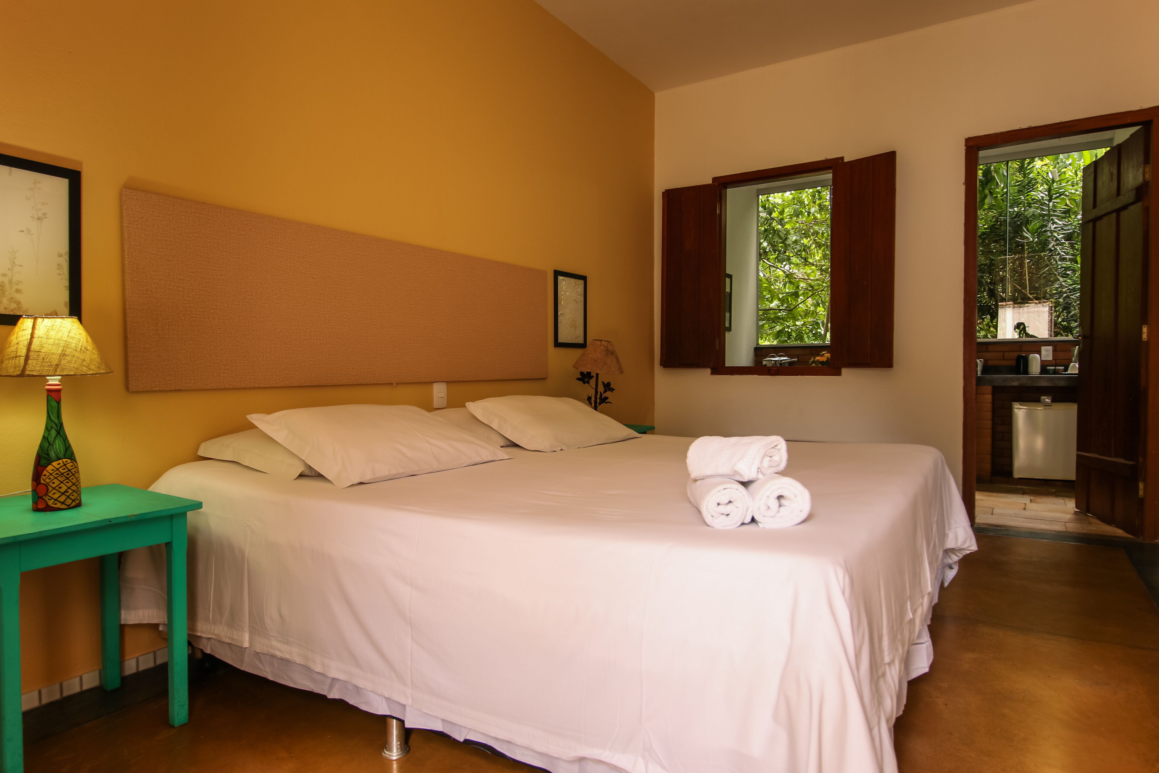 A neatly made bed with white linens and rolled towels, a small table with a lamp, and a window with open shutters.