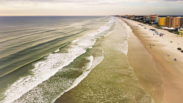 A beach with a clear view of the ocean, a sandy shore, and a row of buildings along the coastline.