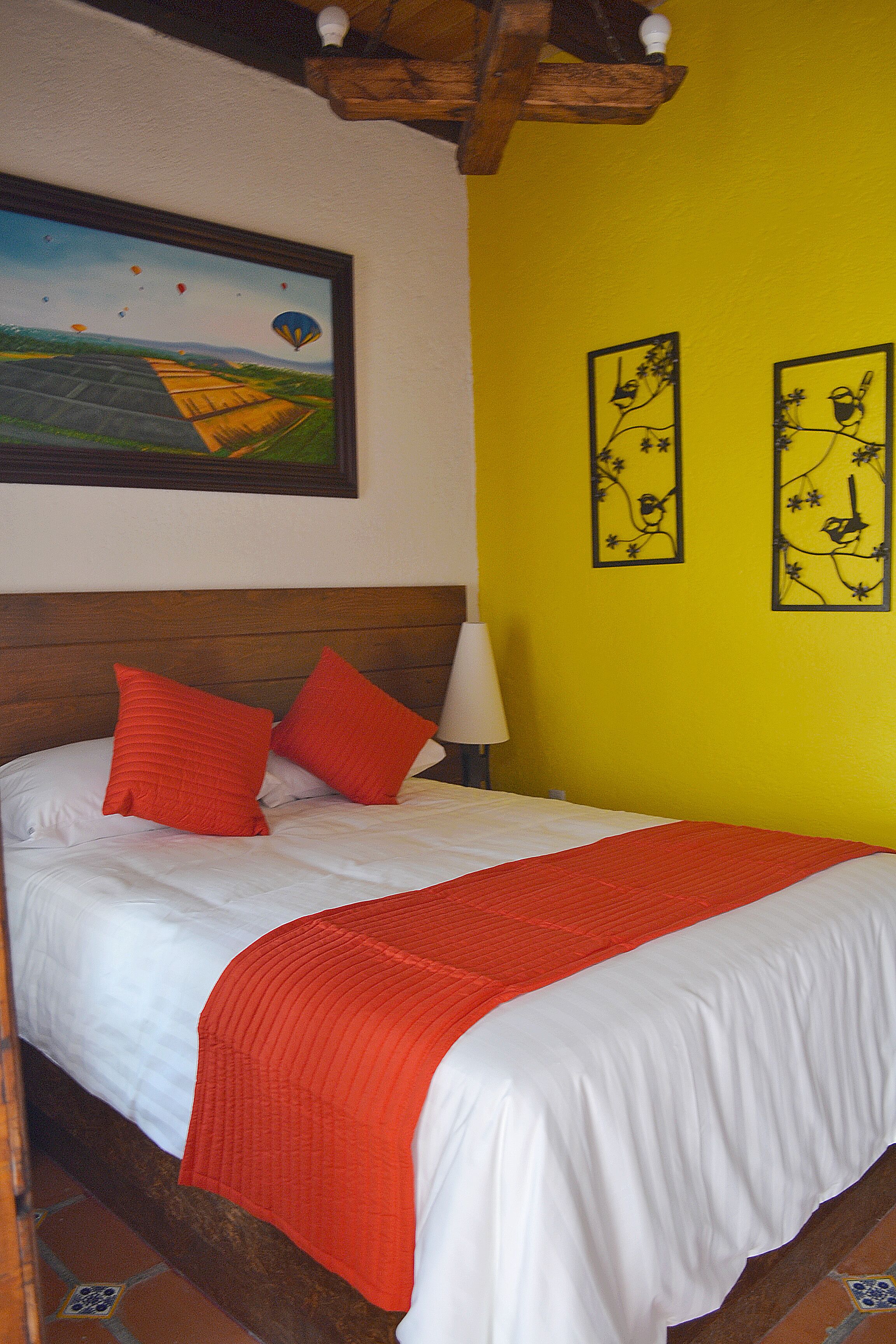 Een bed met witte beddengoed en rode kussens, een houten hoofdbord en een gele muur met twee zwarte decoratieve wandobjecten.