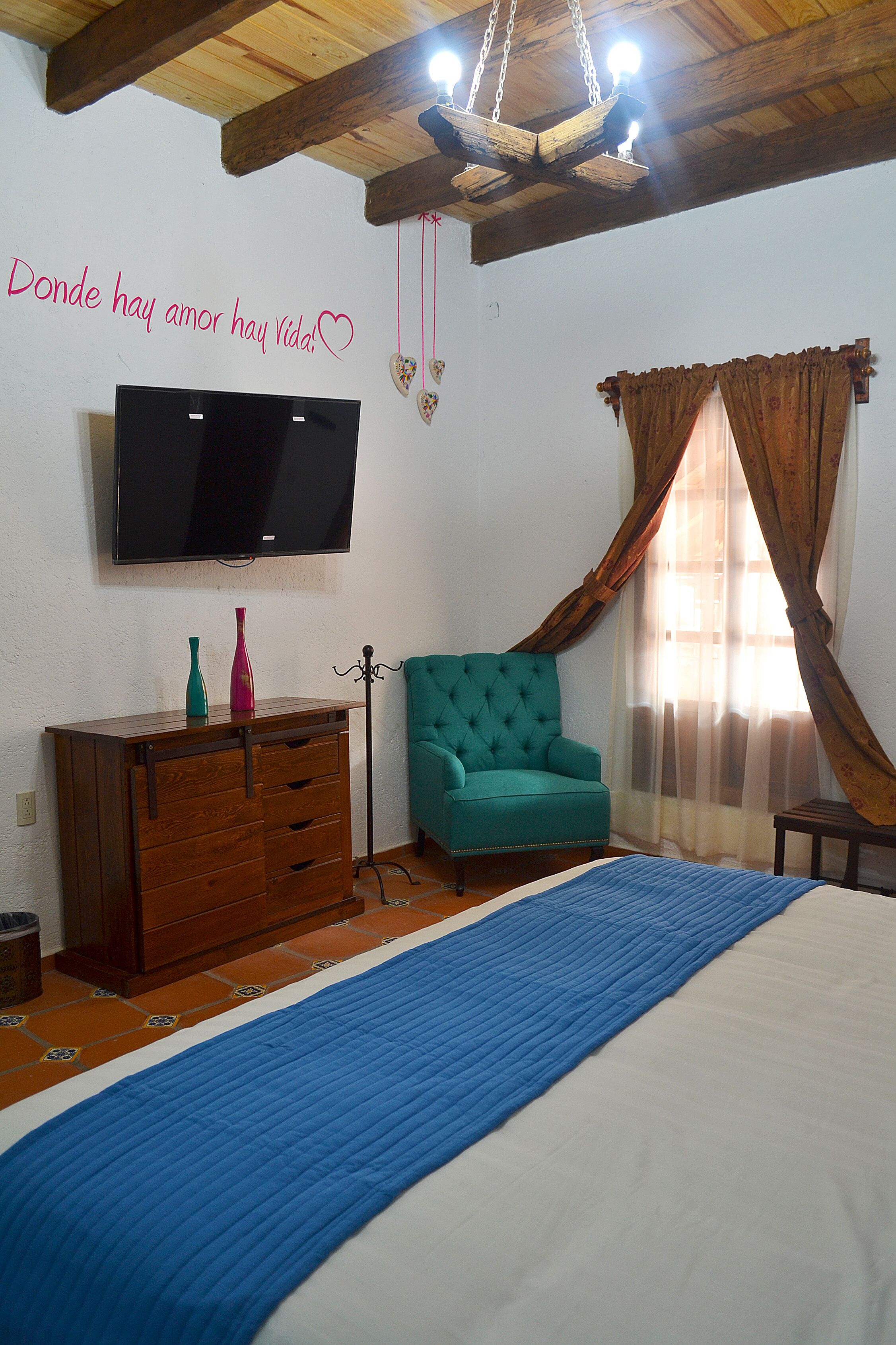 Une chambre à coucher comprenant un lit, une commode en bois, un fauteuil turquoise, un téléviseur fixé au mur et une fenêtre avec des rideaux.