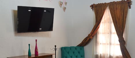 A bedroom with a bed, a wooden dresser, a teal armchair, a TV mounted on the wall, and a window with curtains.