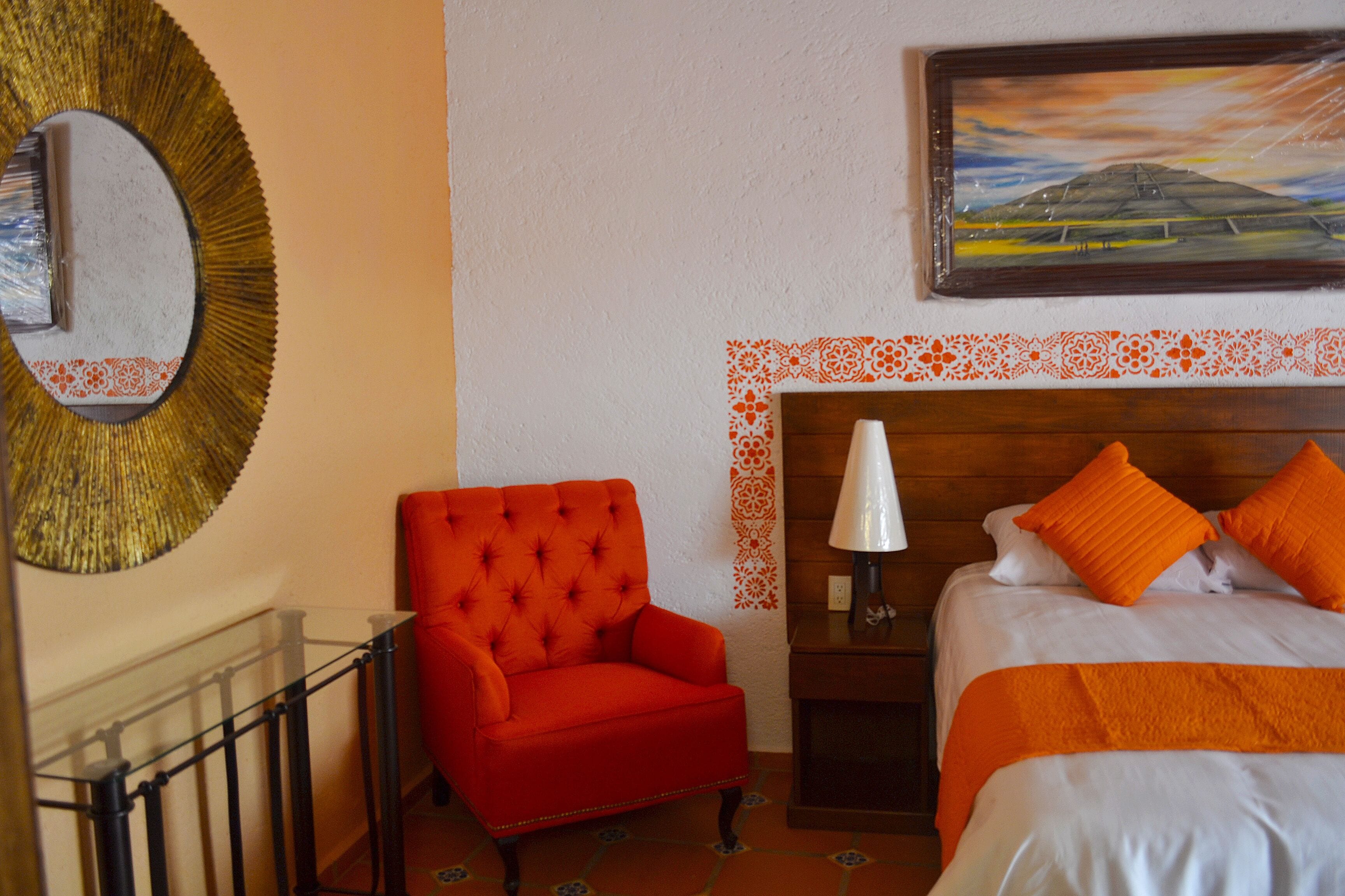 A hotel room with a red armchair, a bed with orange bedding, a wooden headboard, a nightstand with a lamp, and a painting above the bed.
