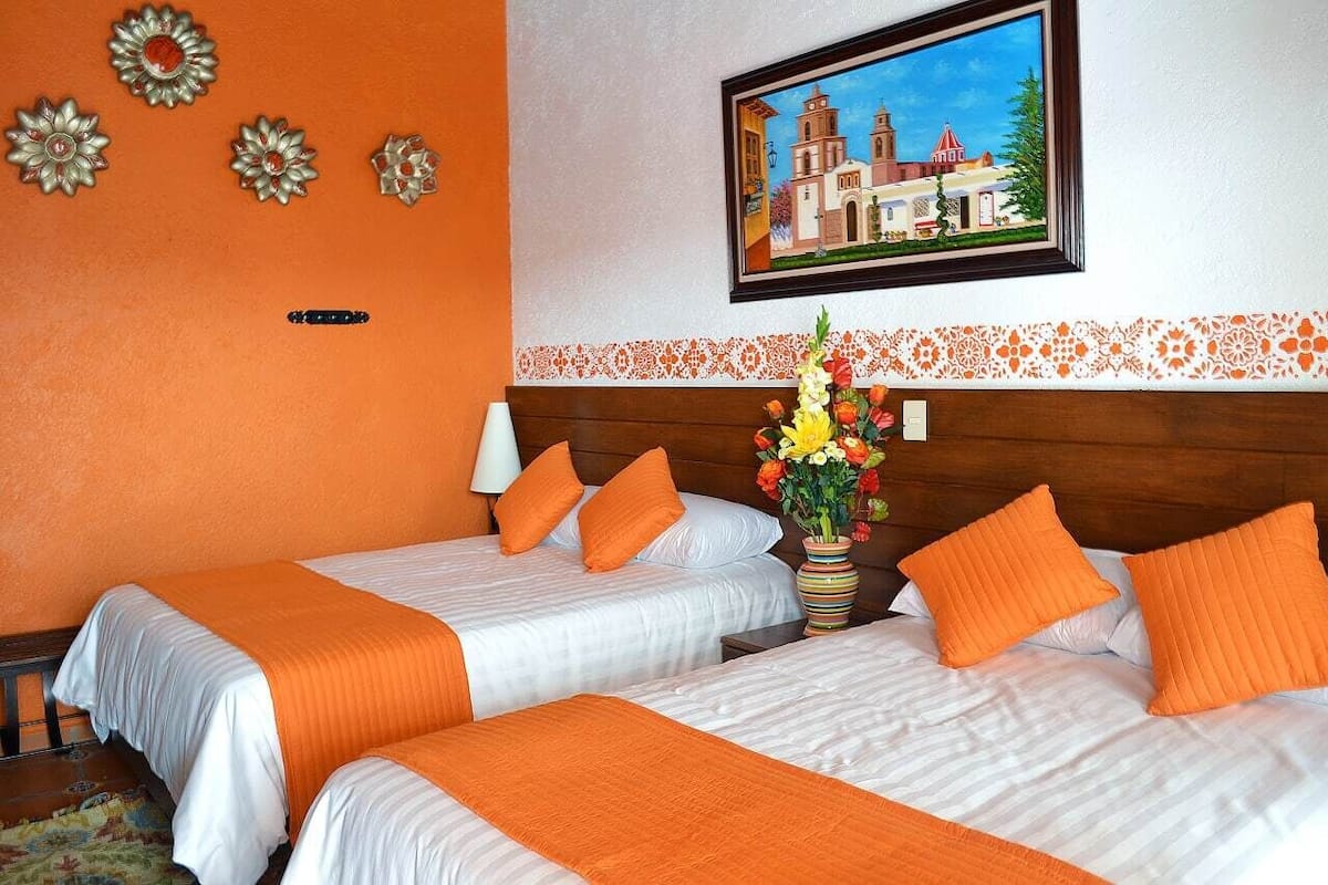 Ein Hotelzimmer mit zwei Betten, orangefarbener Bettwäsche, einem Holz-Kopfteil, einem Bild eines Gebäudes und einer Vase mit Blumen.