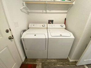 A laundry room with two white front-loading washers and dryers, a wooden shelf above, and a door with a doorknob. - 3BR Family Home | Red Rock Views+Stargazing (Kanab)