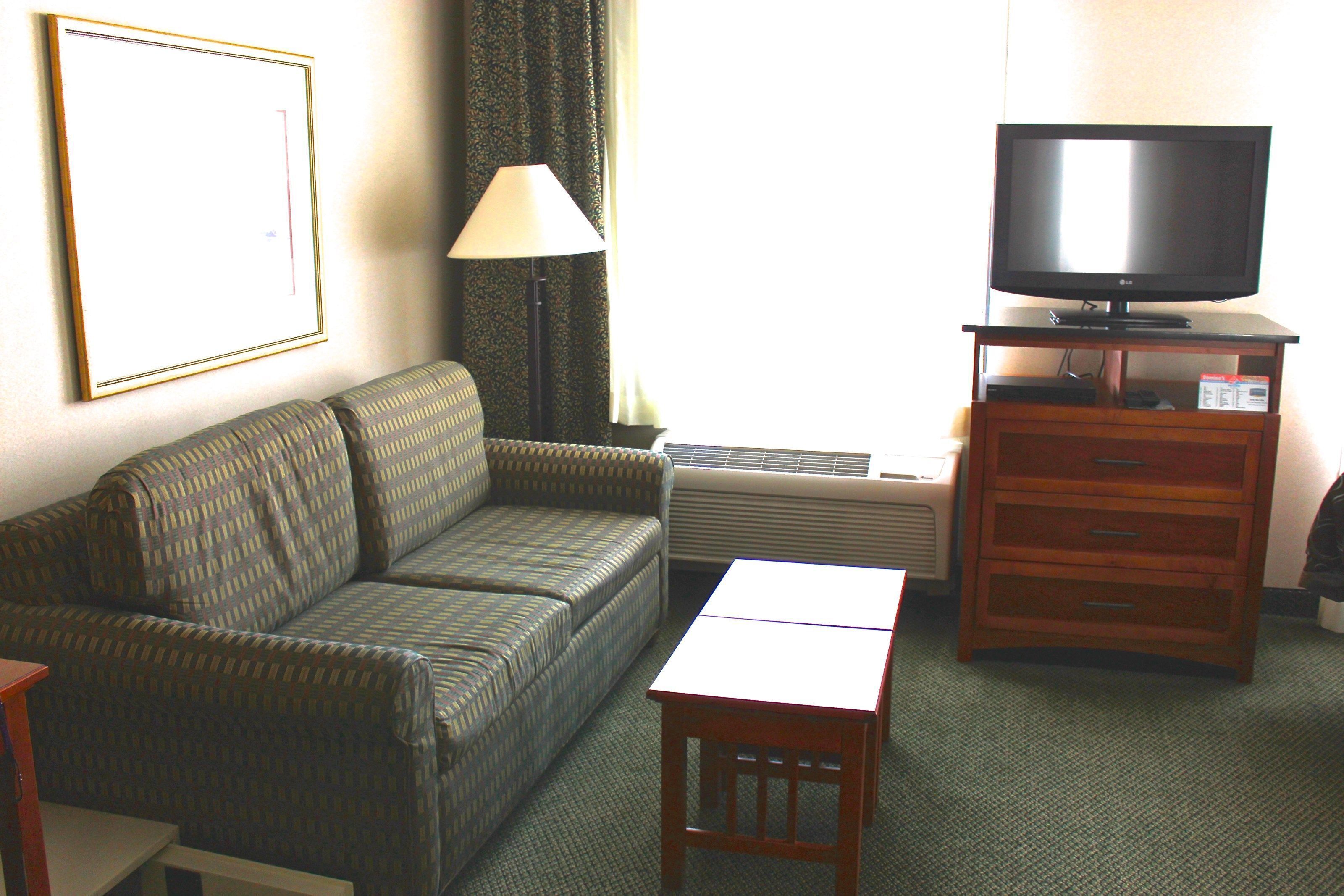 A hotel room with a sofa, a small table, a TV on a wooden stand, and a lamp.