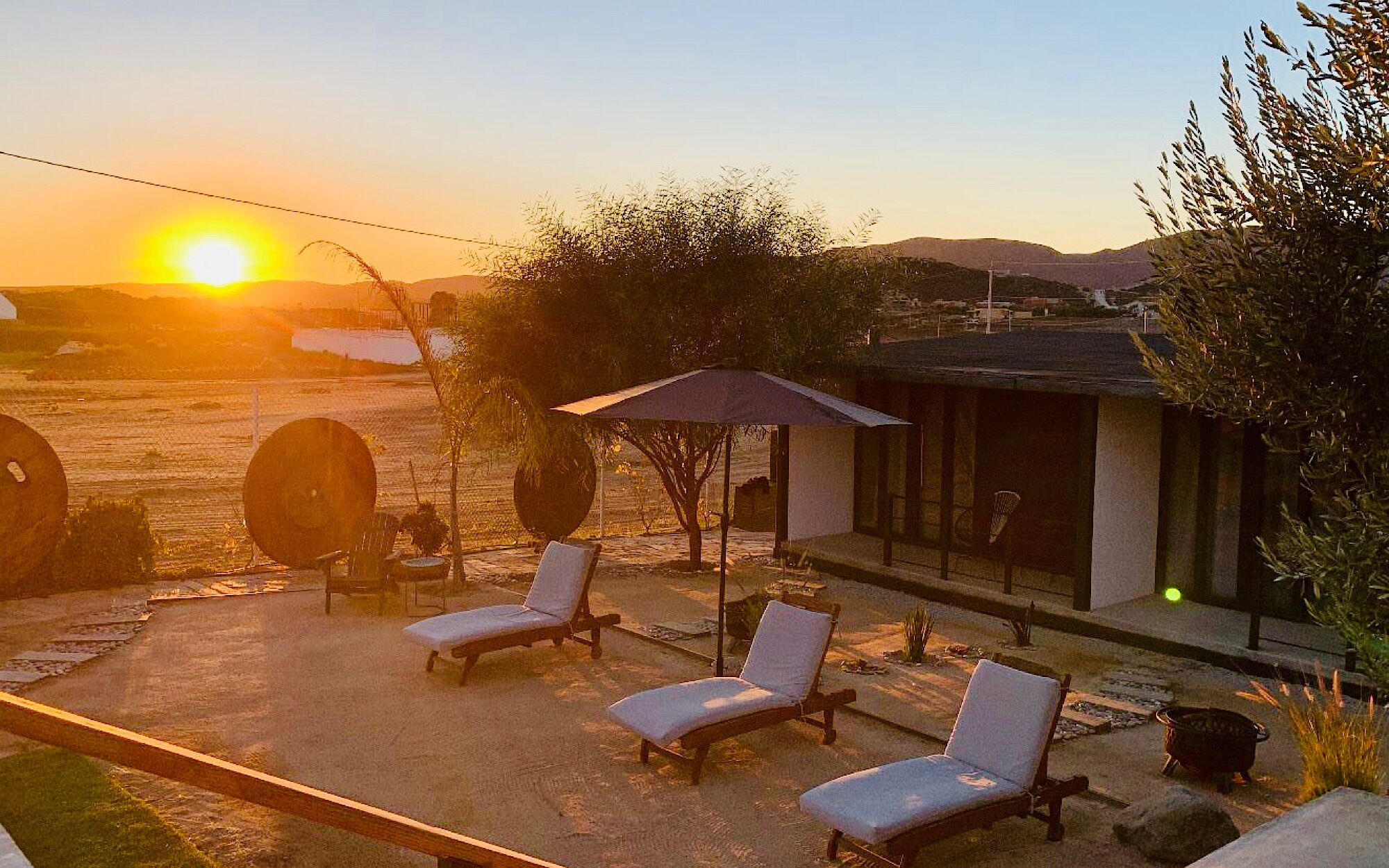 Un patio con sillones de descanso y un área de estar cubierta, con vistas a un paisaje escénico con colinas y un atardecer.
