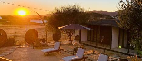 Un patio con sillones de descanso y un área de estar cubierta, con vistas a un paisaje escénico con colinas y un atardecer.