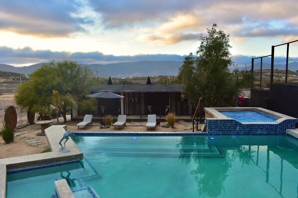 A swimming pool with lounge chairs and a hot tub, surrounded by trees and a view of mountains.