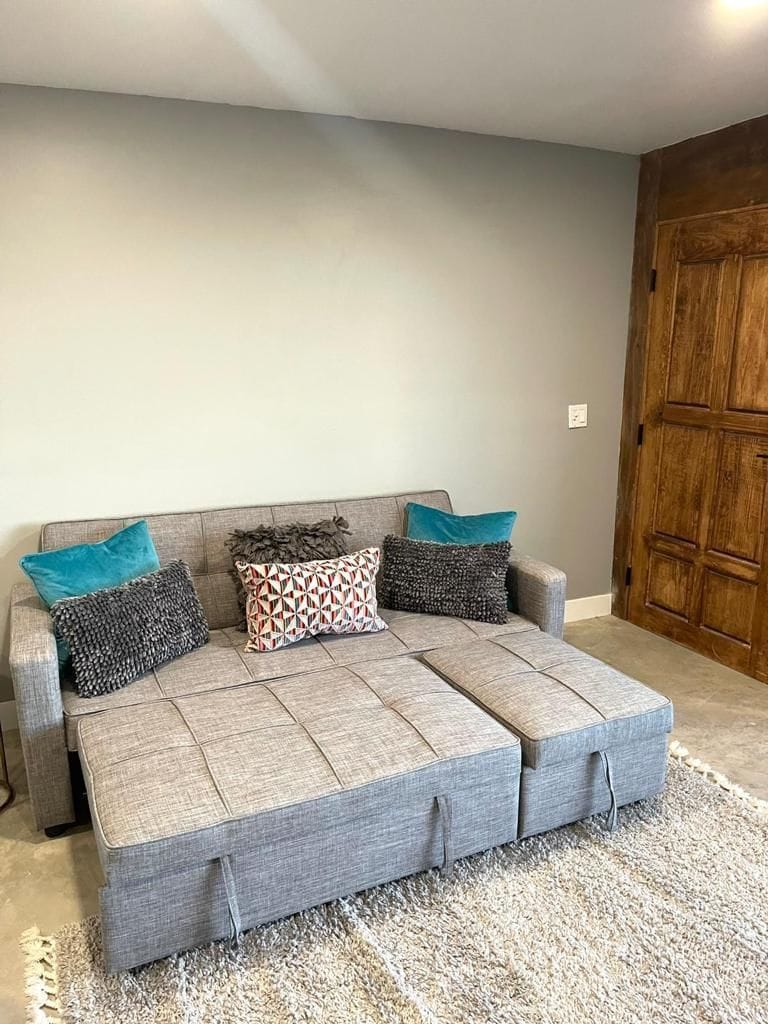 A grey sectional sofa with multiple cushions against a plain wall, a wooden door to the right, and a textured rug on the floor.