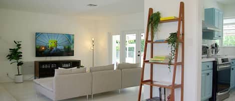 A modern living room with a sofa, a TV mounted on the wall, a bookshelf, and a kitchen area with cabinets and appliances.