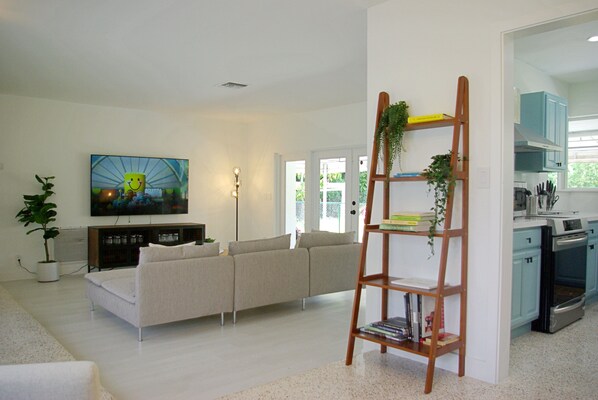 A modern living room with a sofa, a TV mounted on the wall, a bookshelf, and a kitchen area with cabinets and appliances.