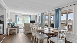 En rummelig spiseplads med et træbord og hvide stole, store vinduer med blå gardiner og udsigt til en balkon.