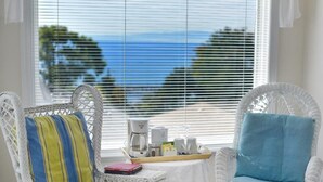 A room with two wicker chairs, one with a striped cushion and the other with a checkered cushion, a small table with a tray of items, and a view of the sea through large windows. - Star of the sea Guest house, Sea Forever King size bed with ocean view balcony! (White Rock)